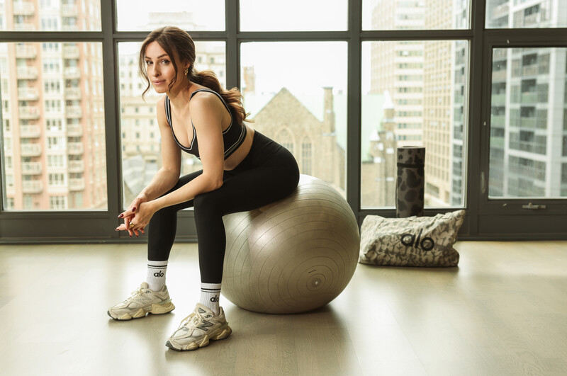 Woman in black athletic wear balanced on a stability ball in a high-rise gym with floor-to-ceiling windows overlooking a city skyline.