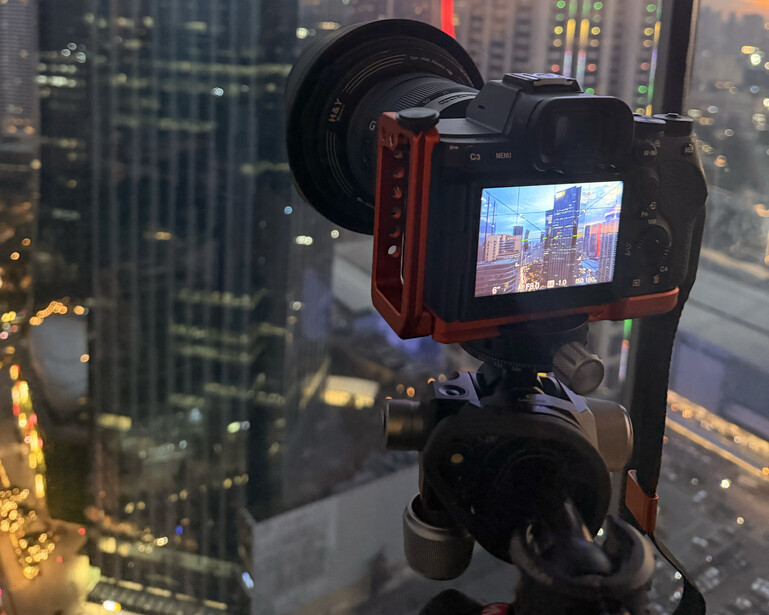 Camera mounted on tripod with articulating red arm, capturing cityscape on screen at dusk.