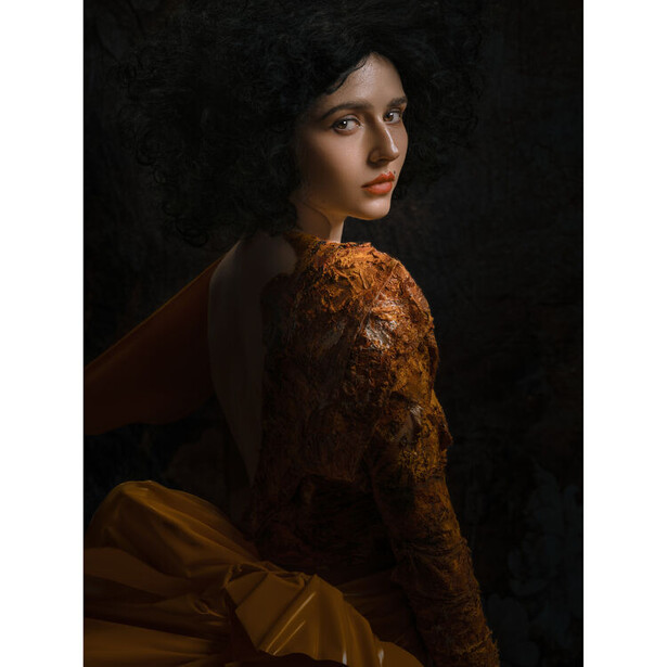 Woman with dark hair wearing rust-colored lace garment, posed against dark background looking over shoulder.