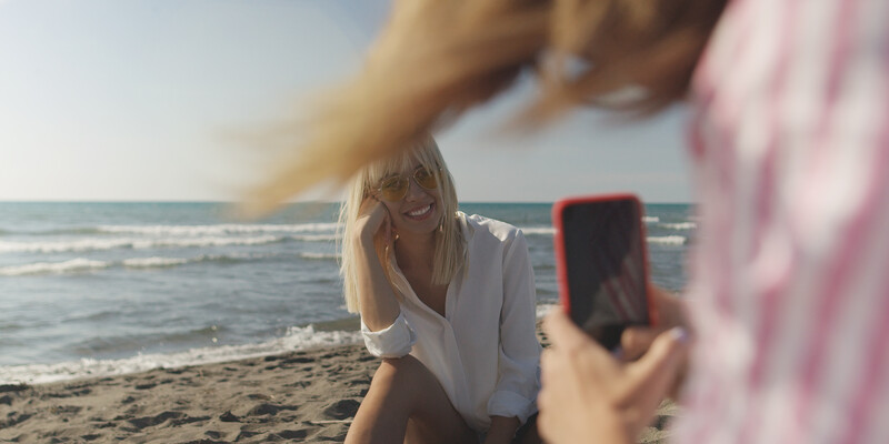 Woman posing for a portrait photo on a beach while someone frames her with a smartphone camera.