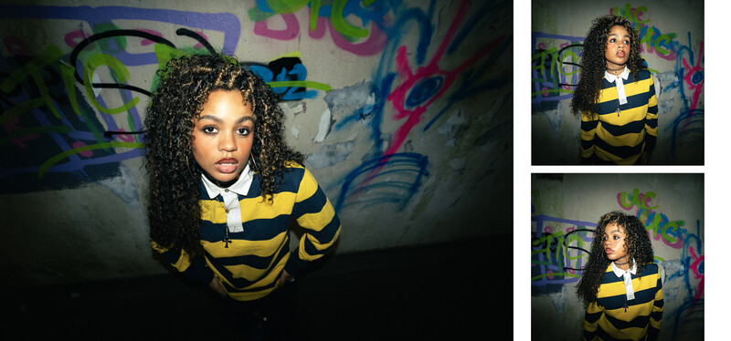 Young woman with curly hair wearing yellow and black striped sweater against colorful graffiti wall.