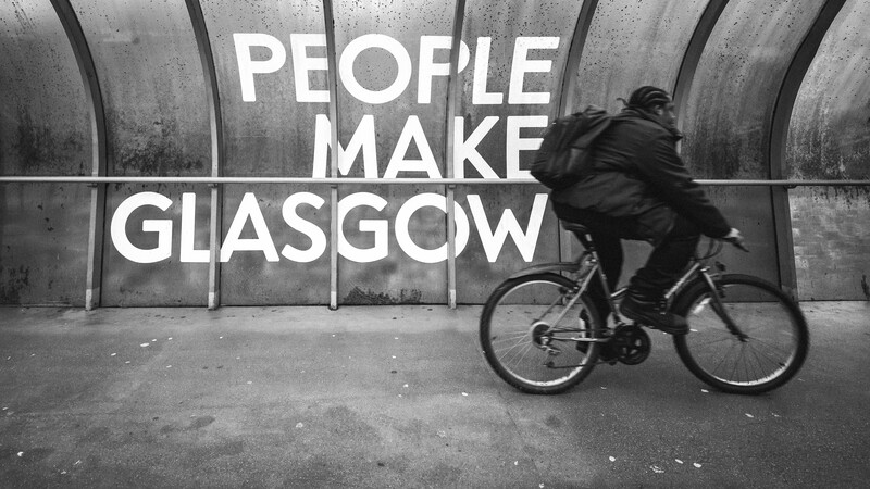 Cyclist riding past concrete wall with white text reading "PEOPLE MAKE GLASGOW".