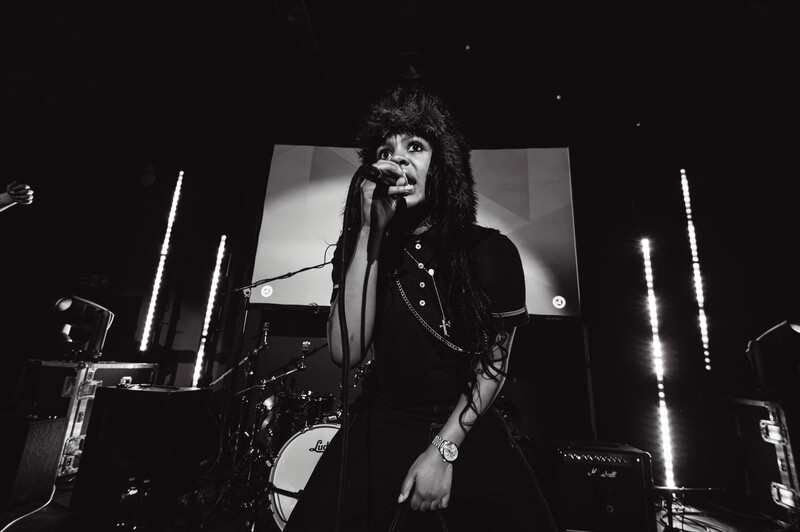 Black and white photograph of a performer singing into a microphone on stage with dramatic vertical lighting.