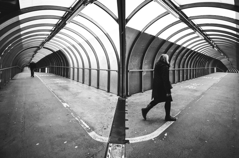 Figure in dark coat walking through symmetrical tunnel with arched ribbed ceiling in black and white.