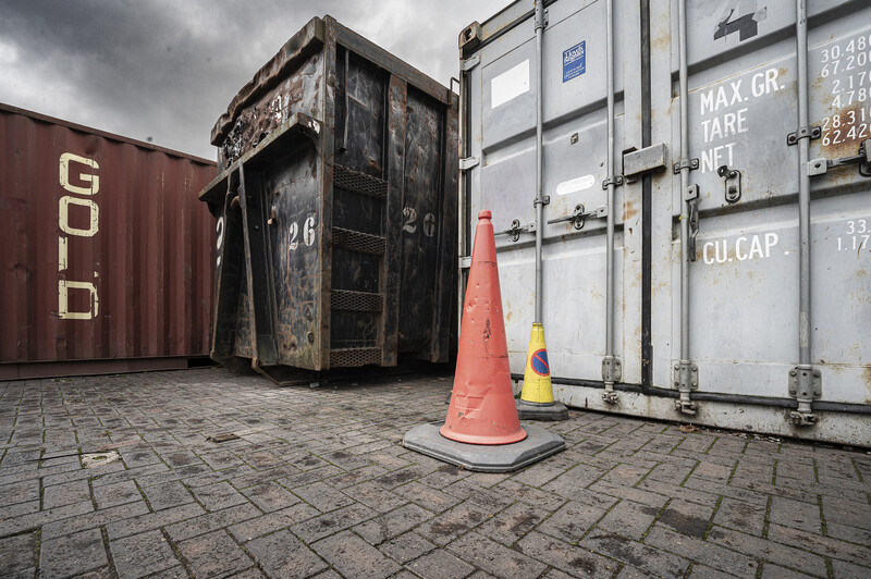 Industrial shipping containers with weathered surfaces and an orange traffic cone on pavement beneath overcast sky.