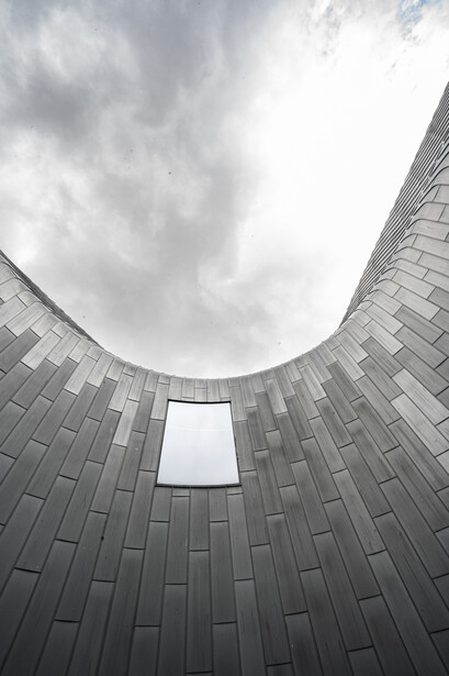 Upward view of a modern curved building facade with gray tiles framing a bright sky opening.
