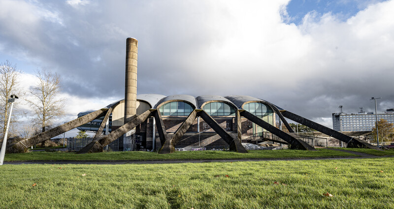 Modernist building with curved steel framework and large circular windows beside a tall chimney, photographed against a cloudy sky.