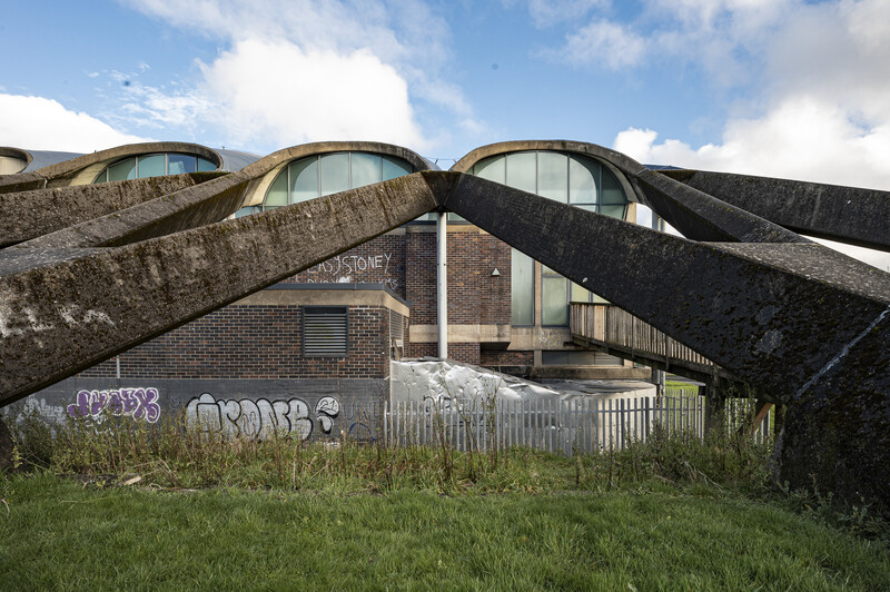 Modernist brick building with three curved concrete roof forms and large windows, surrounded by overgrown grass and bare tree branches.