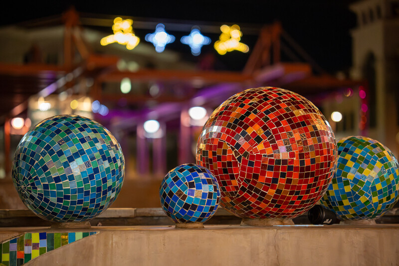 Colorful mosaic-tiled decorative spheres of varying sizes arranged on a ledge with blurred festive lights in the background.