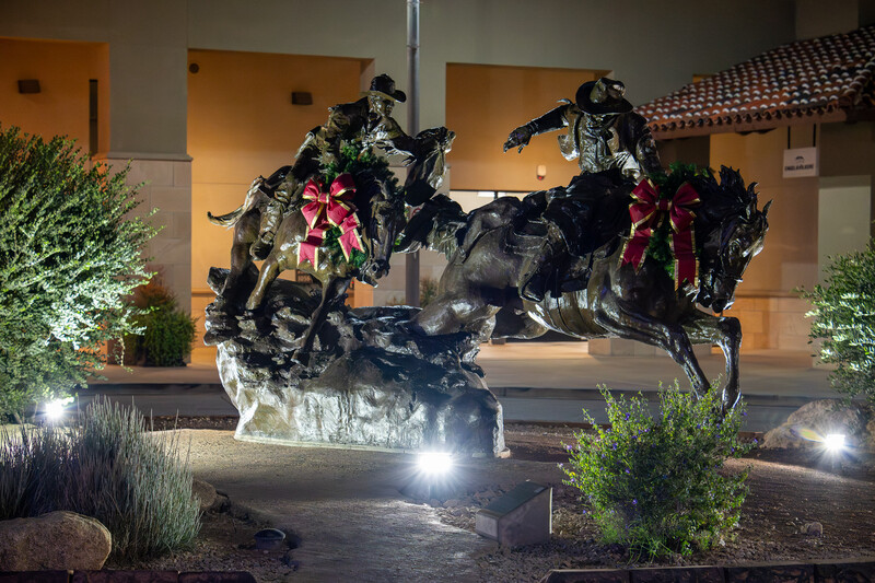 Nighttime photograph of an ornamental metal sculpture featuring two horses with decorative red bows, illuminated by ground-level lighting.