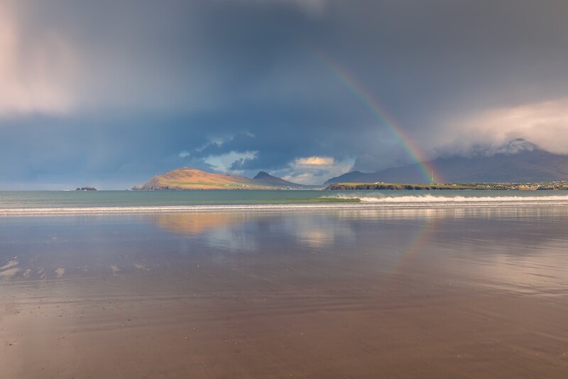 Rainbow arcing over a coastal landscape with wet sand beach, distant mountains, and dramatic cloud formations at sunset.