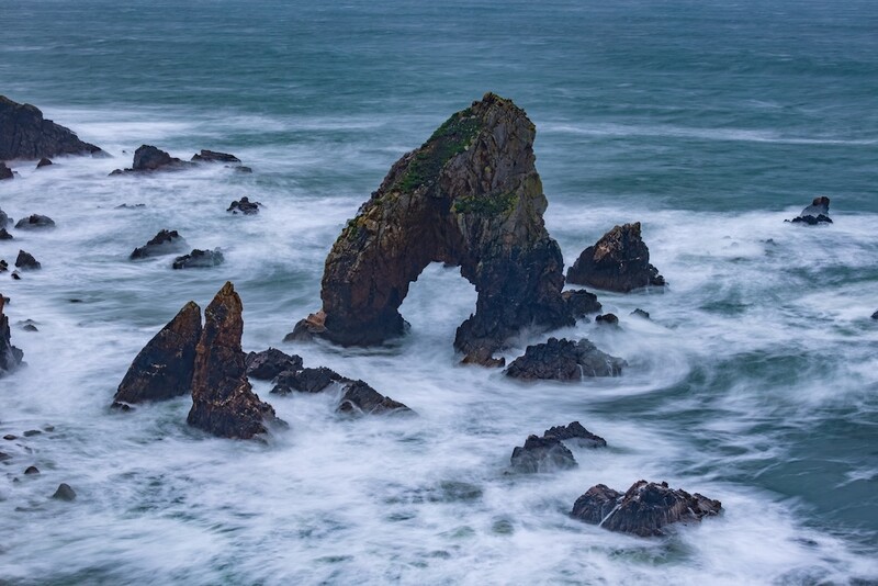 Rock formations with natural arch surrounded by white-capped waves crashing against a rocky coastline.