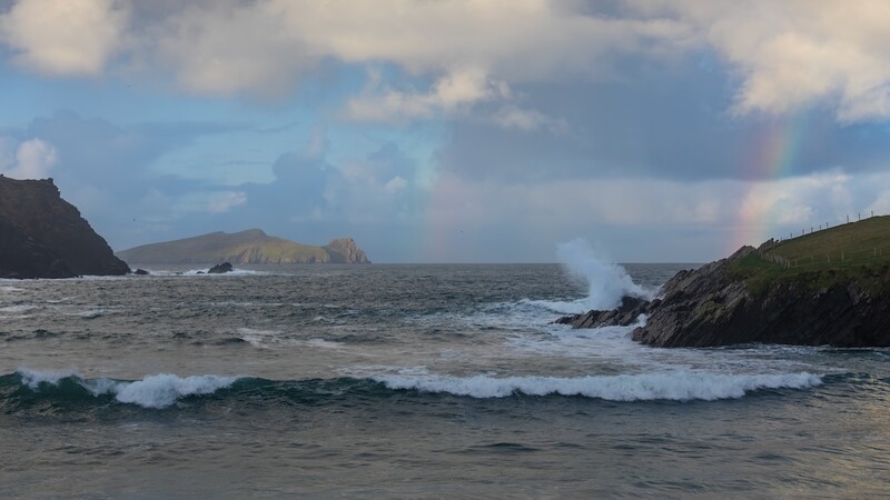 Dramatic coastal landscape with waves crashing against rocky cliffs under partly cloudy sky.
