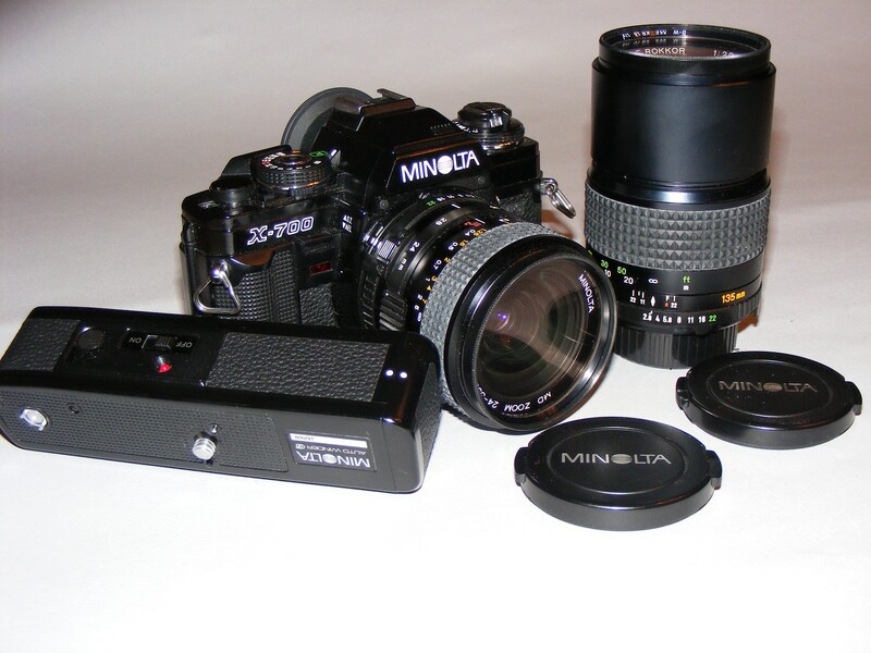 Minolta X-700 film camera with two lenses, lens caps, and battery pack on white background.