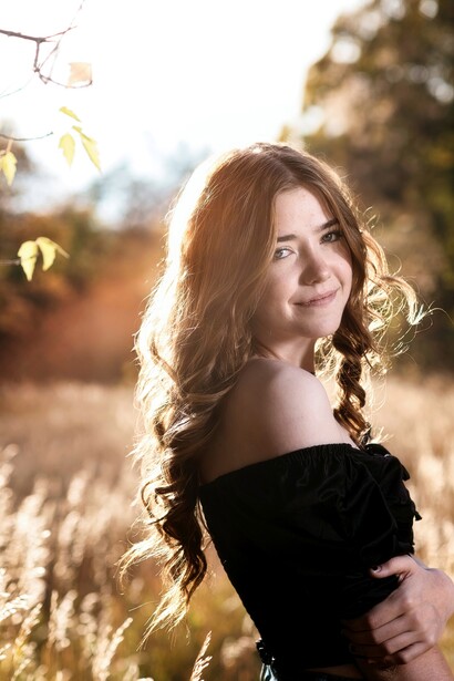 Woman with long wavy hair in off-shoulder black top, backlit by golden hour sunlight in an outdoor setting.