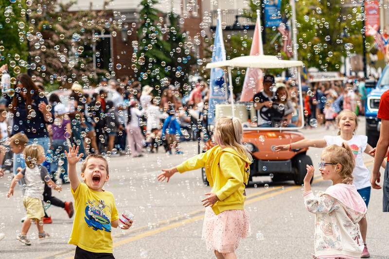 Children in yellow and pink clothing playing among soap bubbles at an outdoor street festival.