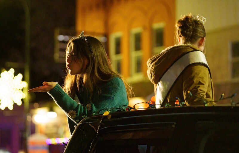 Two young people sitting on a railing at night, backlit by warm city lights and building facades.