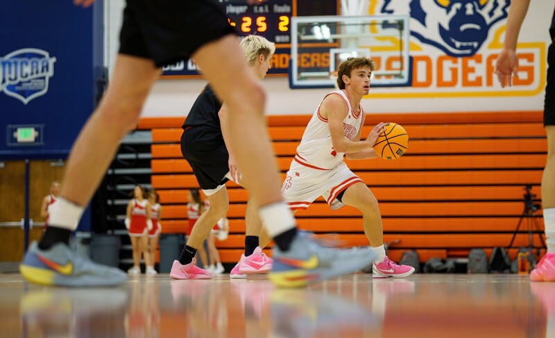 Young basketball player in white jersey dribbling during practice on indoor court with orange bleachers.