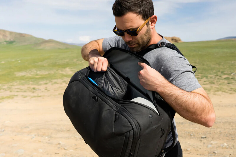 Man wearing sunglasses and gray shirt demonstrates opening a black camera backpack outdoors in rolling hills.
