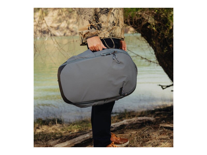 Person in camouflage jacket holding a gray camera bag against a blurred water and trees background.