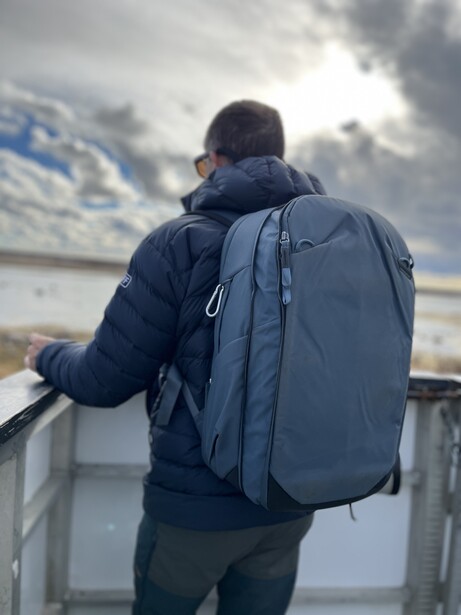 Person wearing a blue backpack and navy jacket standing on an elevated platform overlooking a body of water.