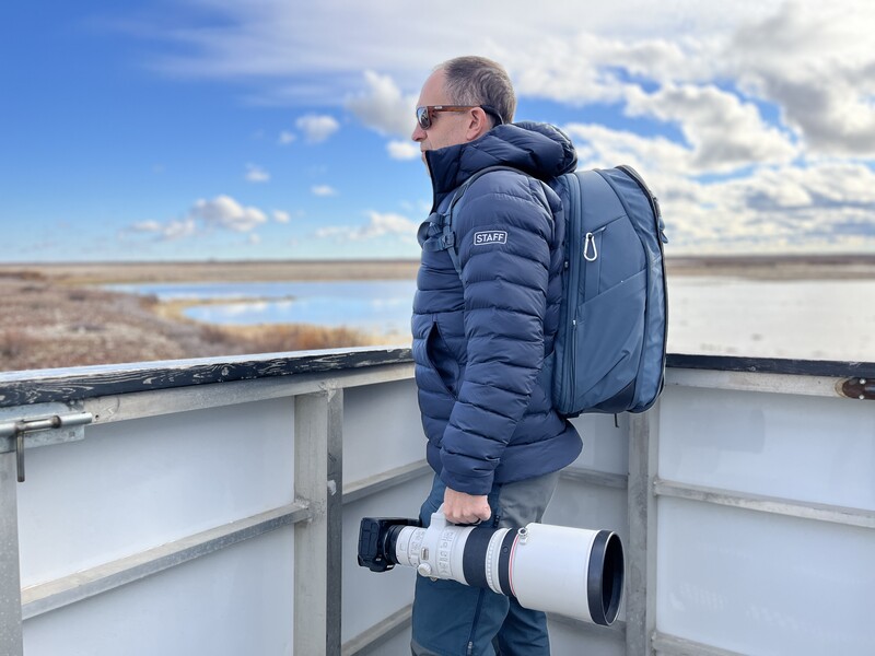 Photographer holding telephoto lens while standing on observation platform overlooking wetland landscape.