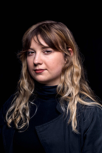 Woman with long wavy blonde hair and bangs wearing a dark turtleneck against a black background.