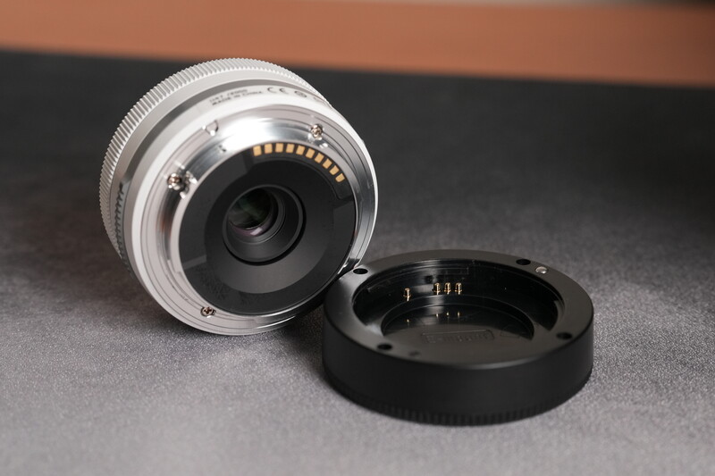 Silver mirrorless lens with detached black rear cap showing electronic contacts.