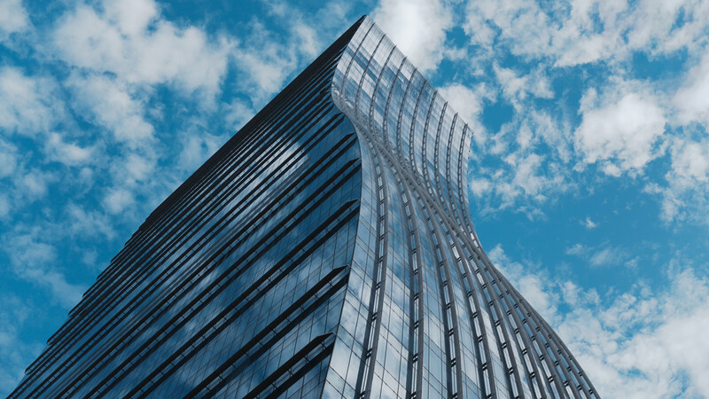 Modern skyscraper with curved glass and metal facade photographed from below against blue sky with white clouds.