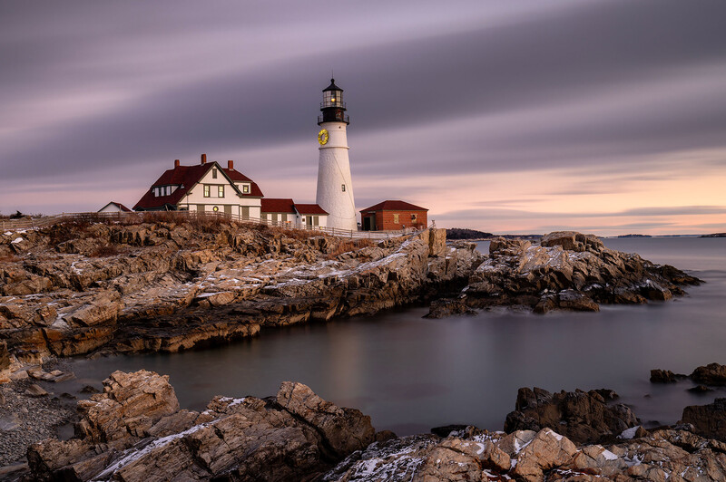 White lighthouse with red roof on rocky coastal peninsula at dusk with keeper's house nearby.