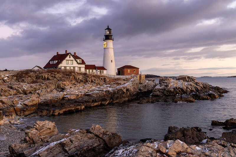White lighthouse with black top on rocky coastal peninsula beside residential buildings at dusk.