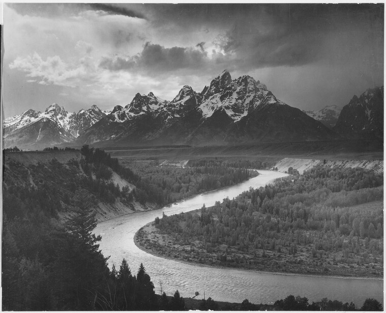 Mountain range with dramatic peaks reflected in a winding river below, surrounded by forested valleys under stormy skies.
