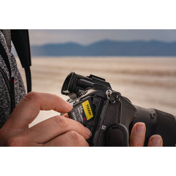 Photographer holding a DSLR camera with hands in frame, beach and distant mountains visible in background.