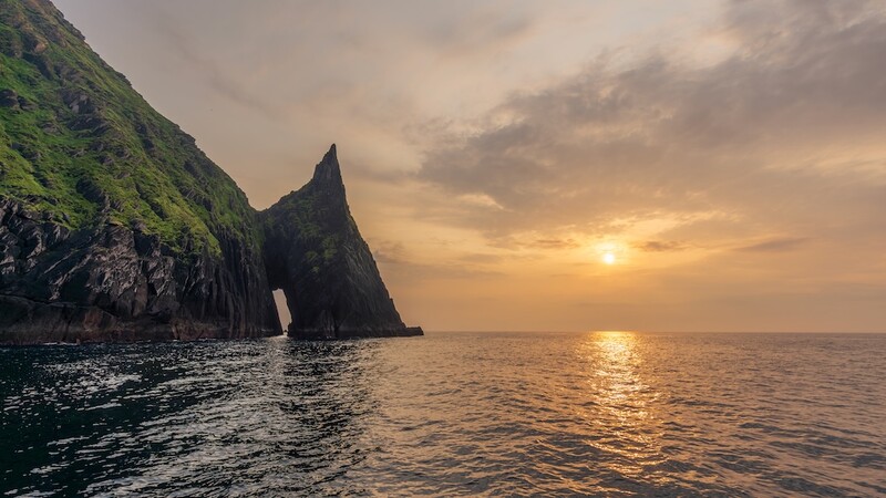 Dramatic sea arch with moss-covered cliff face at golden hour sunset over calm ocean.