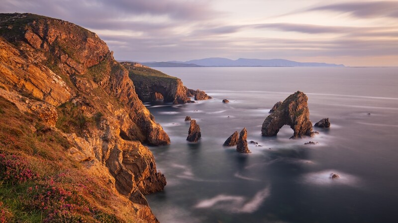 Rocky coastal cliffs at golden hour with sea stacks and smooth water from long exposure.