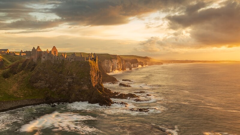 Medieval castle ruins perched on a clifftop overlooking a dramatic coastline at sunset.