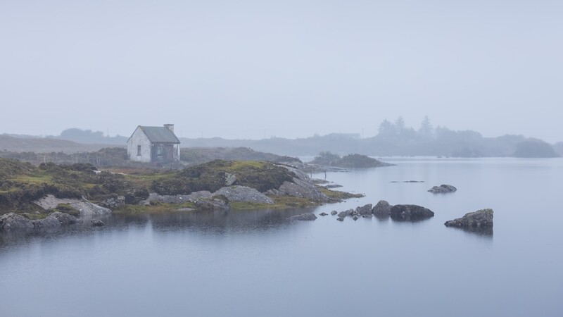 Solitary white cottage on rocky shoreline surrounded by calm water and distant forested islands in heavy fog.