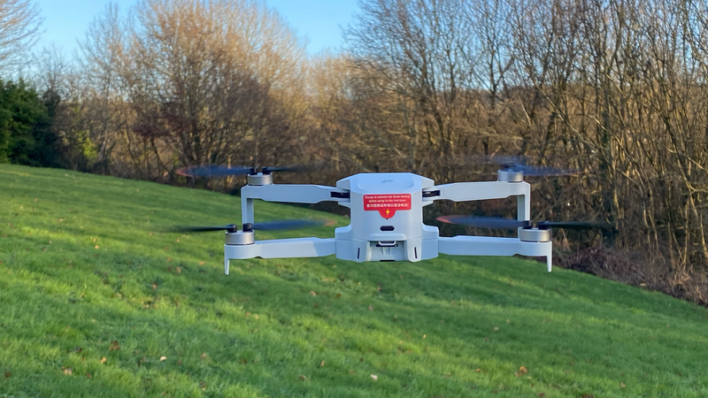 White compact drone with folded arms positioned on grass with bare trees in background.