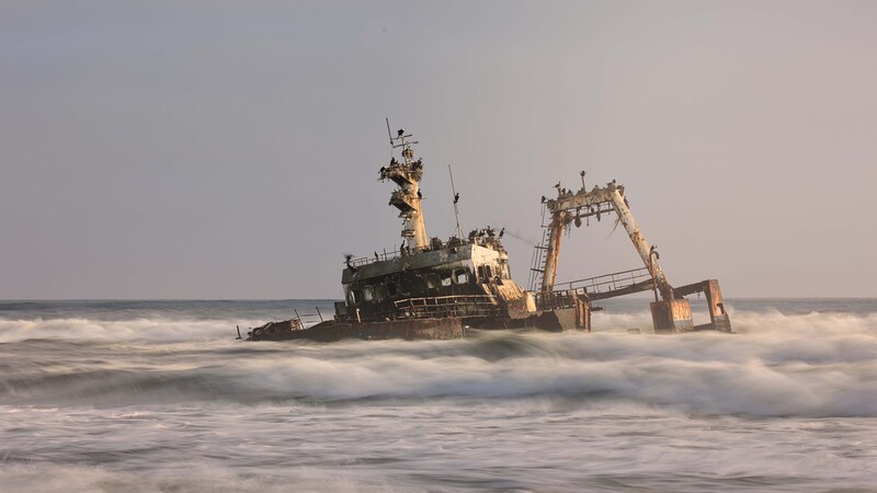 Abandoned cargo ship listing in shallow ocean waves at sunset.