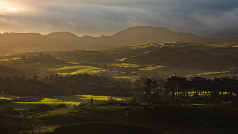 Rolling countryside landscape with golden fields beneath dramatic clouded sky and distant mountains.