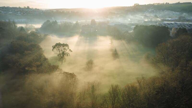Misty valley landscape at sunrise with trees emerging through dense fog and warm light breaking through clouds.