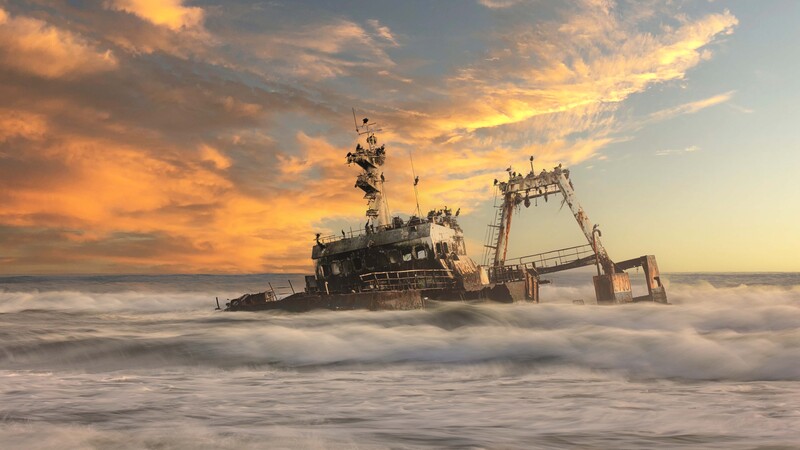 Abandoned ship wreck partially submerged in ocean waves at golden hour sunset.