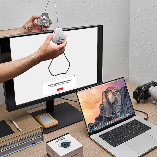 Hands holding a wireless computer mouse above a desk with a monitor, laptop, and photography equipment.