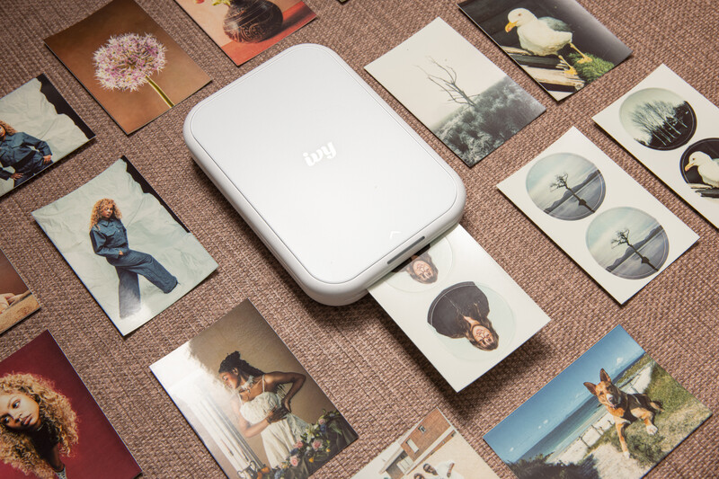 White portable photo printer surrounded by printed photographs arranged on a textured beige surface.