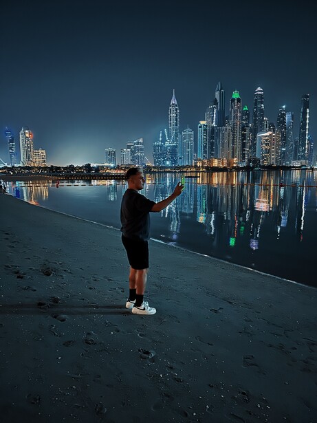 Silhouetted figure standing on a beach at dusk, facing a brightly lit city skyline reflected in still water.