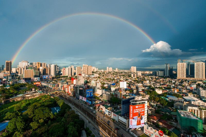 A complete double rainbow arcs over a sprawling urban cityscape with high-rise buildings and residential neighborhoods under dramatic storm clouds.