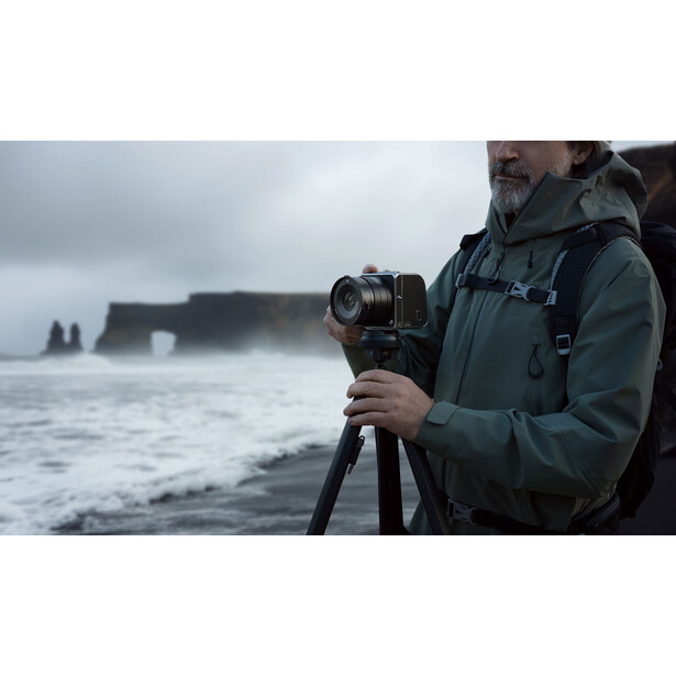 Photographer holding a camera on a tripod while facing a dramatic rocky coastline under overcast skies.