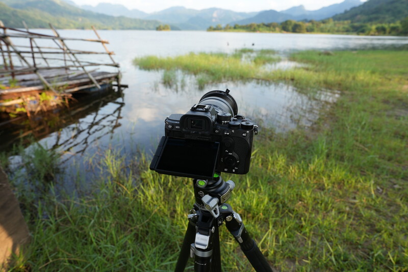 DSLR camera on tripod positioned in shallow water with mountains and wetlands in background.