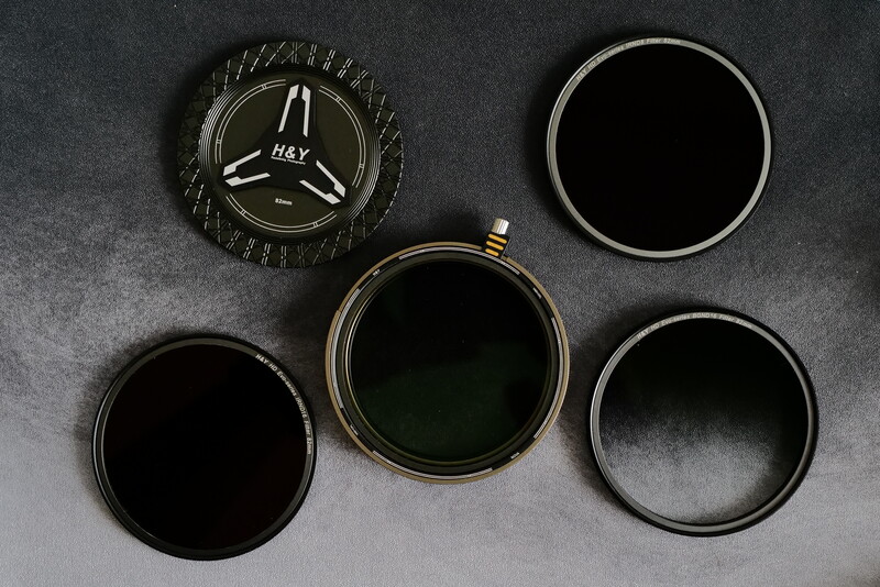 Five camera lens filters and circular glass elements arranged on gray surface, including one marked HAY.