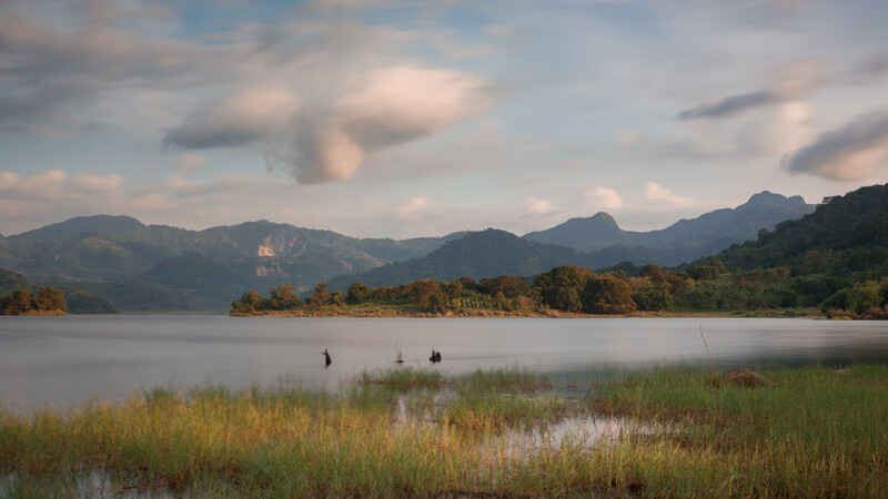 Misty lake landscape with grass-covered shoreline, distant mountains, and three figures wading in shallow water.